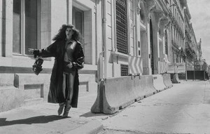 Model Paola Manes walking along a city street sidewalk in a long coat, miniskirt, and slouchy boots beside concrete barriers.