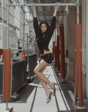 Model hanging from scaffolding pipes on an urban street wearing a black athletic outfit and heeled sandals