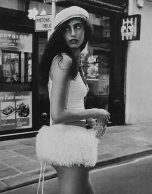 Black and white portrait of a woman wearing a white beret and feathered skirt walking on a city street.