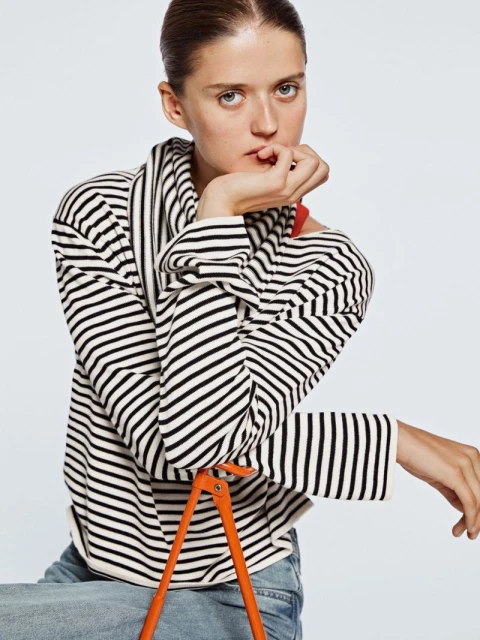 Portrait of a fair-skinned woman in a striped sweater and blue jeans posing in a light studio against a seamless white backdrop.