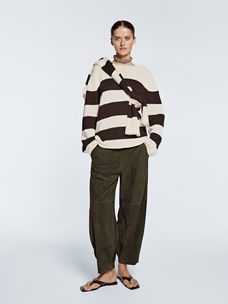 Model wearing a black and off-white striped knit sweater, draped knitwear, dark olive trousers, and flat sandals on a pale background.