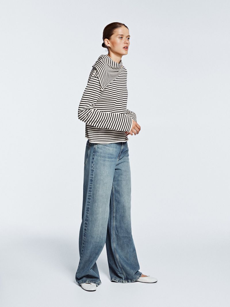 Fashion shot of a woman posing in a striped top and wide-leg denim jeans in a studio setting with a neutral background and diffused lighting.