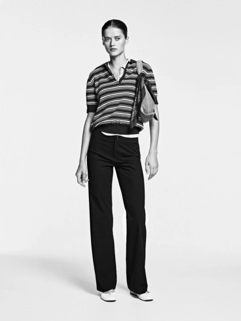 Full-length studio portrait of a woman in a striped top and dark pants, accented with a denim bag, against a bright neutral background.
