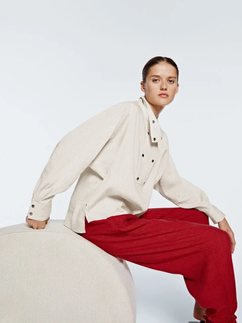 A woman in a light-colored striped shirt and red pants sits on a cream-colored round ottoman in a brightly lit photo studio.