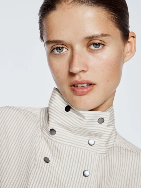 Close-up portrait of a young adult woman with light skin and blue eyes, wearing a striped jacket with metal buttons, on a light background.