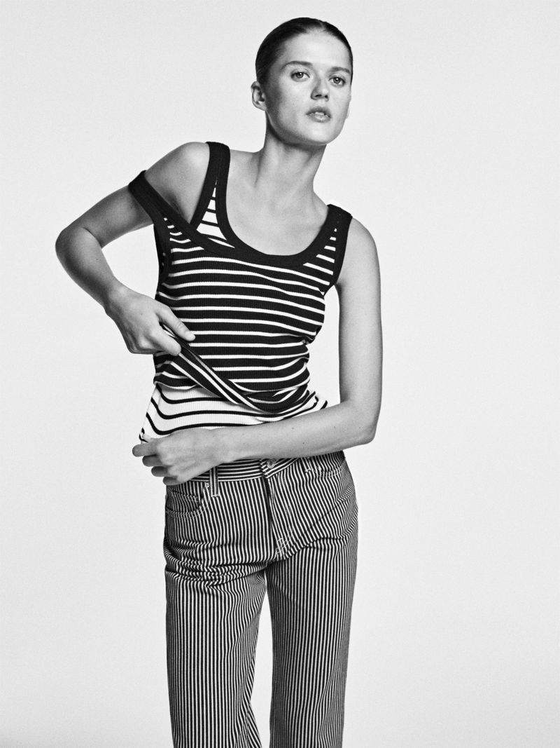 Black and white portrait of a model in a striped top and pants, adjusting her strap in a photo studio against a white background.