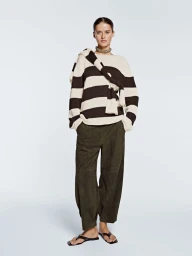 Model wearing a black and off-white striped knit sweater, draped knitwear, dark olive trousers, and flat sandals on a pale background.
