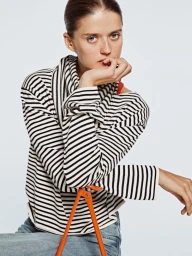 Model wearing a black and white striped top sitting on an orange chair against a plain white background