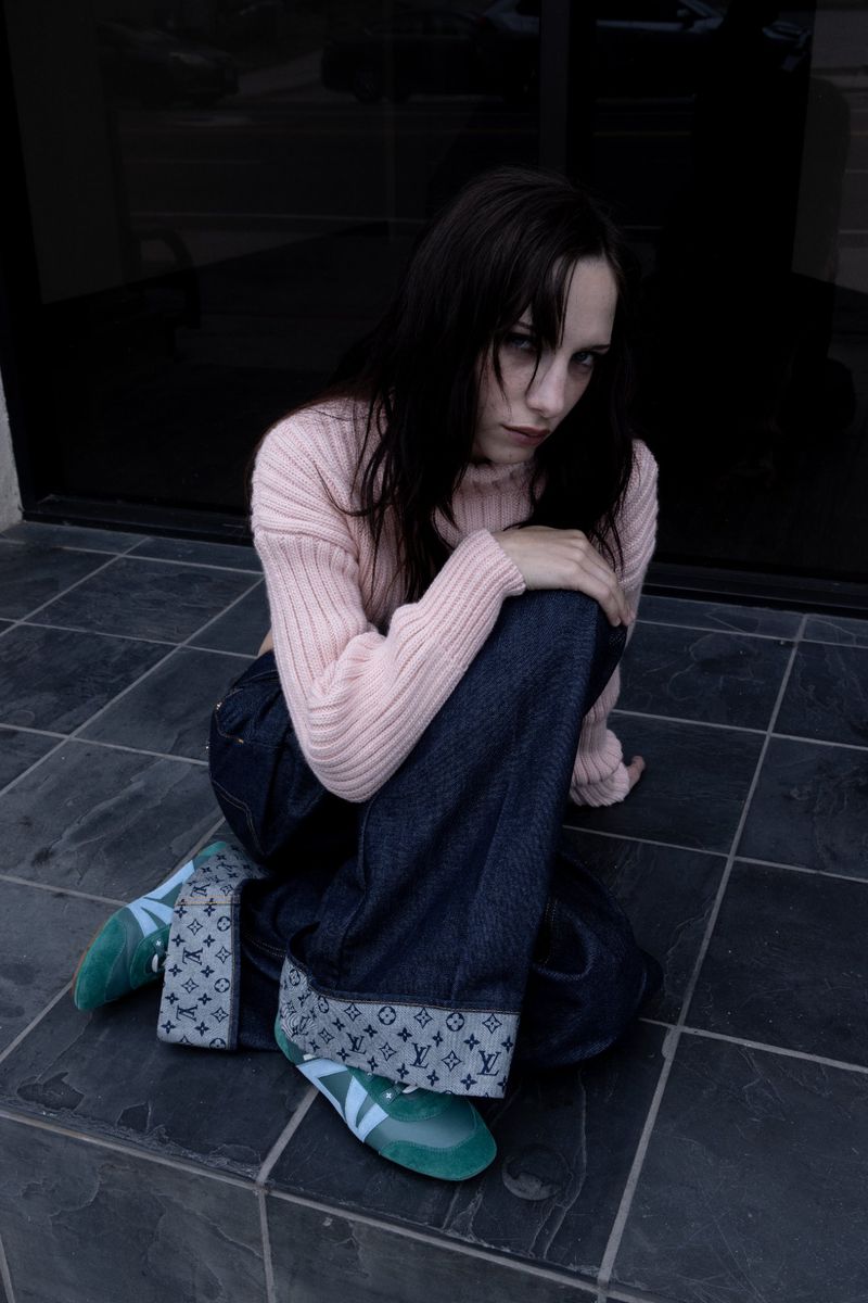 Young woman in a pink sweater and wide-leg jeans sitting on gray tile. She is wearing monogrammed sneakers with green soles.