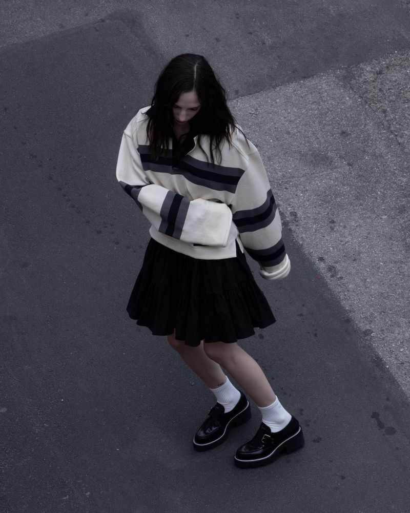 Woman in striped sweater and black skirt walking on gray pavement, viewed from a high angle, with black shoes and white socks.