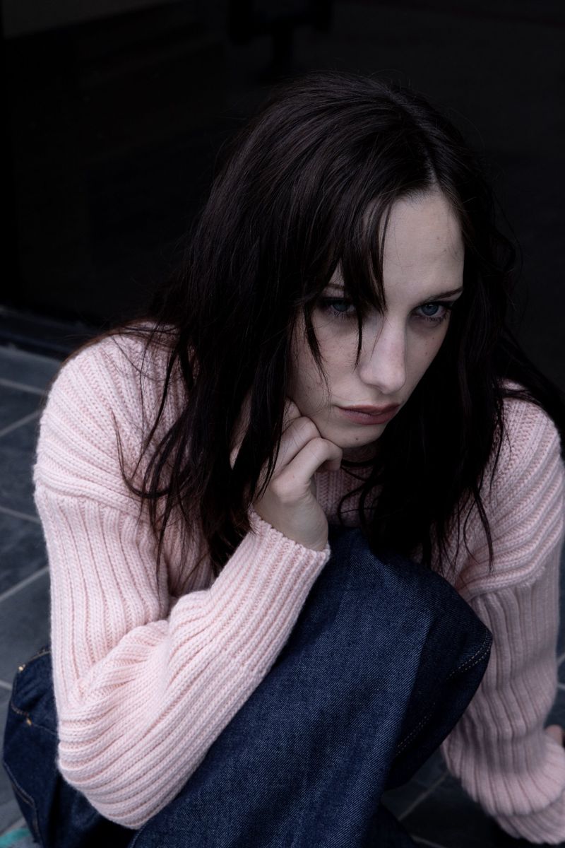 Portrait of a young woman with dark hair and blue eyes wearing a pink sweater and blue jeans, seated against a dark backdrop.