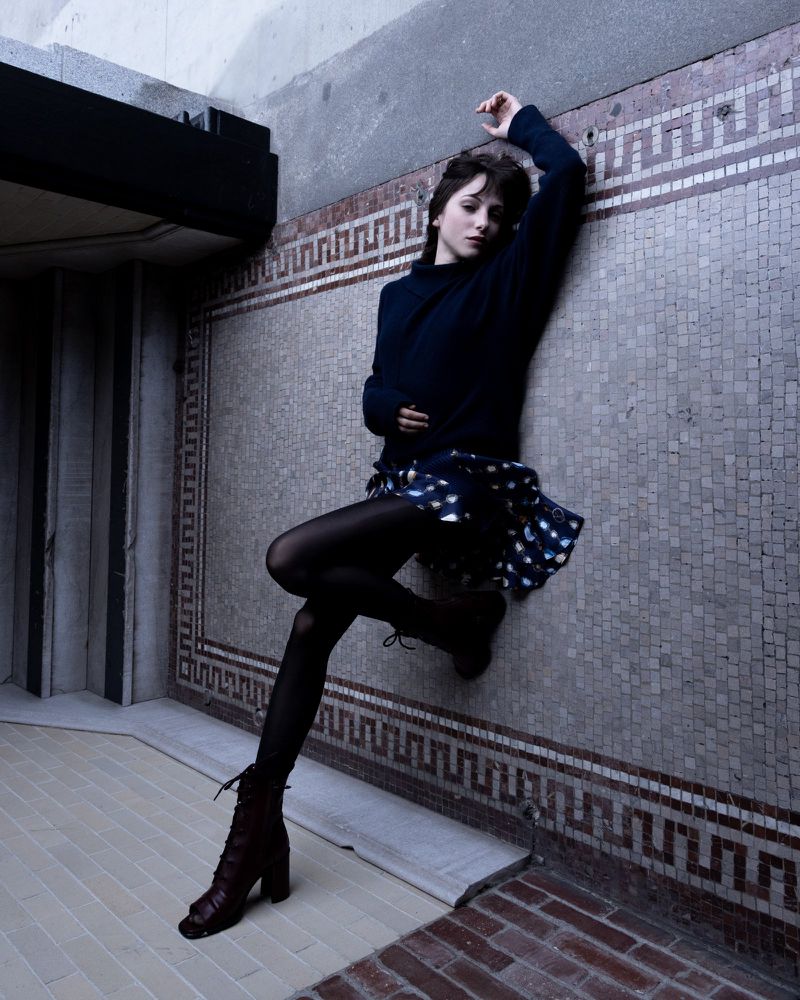 Stylish woman in dark top and patterned mini skirt poses on tiled wall, wearing black tights and lace-up heeled boots in an urban setting.
