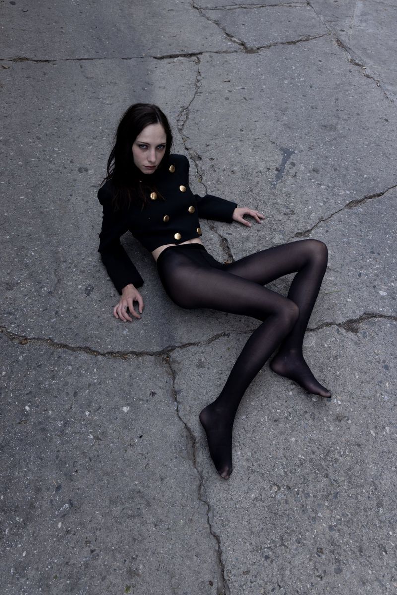 Woman in black jacket and tights lies on cracked concrete. Dark hair and eyes create a striking fashion portrait in muted tones.