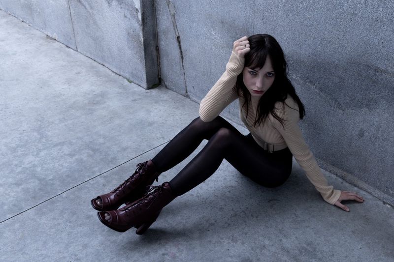 A young woman with dark hair, wearing a tan sweater and black tights, sits on a gray surface with vintage-style open-toe heels.