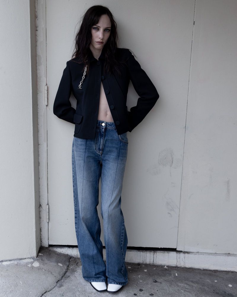 A woman in a black blazer and blue jeans poses against a grey wall, showcasing a fashion-forward and stylish look in an outdoor setting.