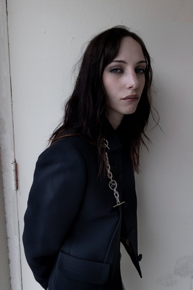 Portrait of an adult woman with long dark hair, wearing a navy blazer with a decorative silver chain on a neutral-colored background.