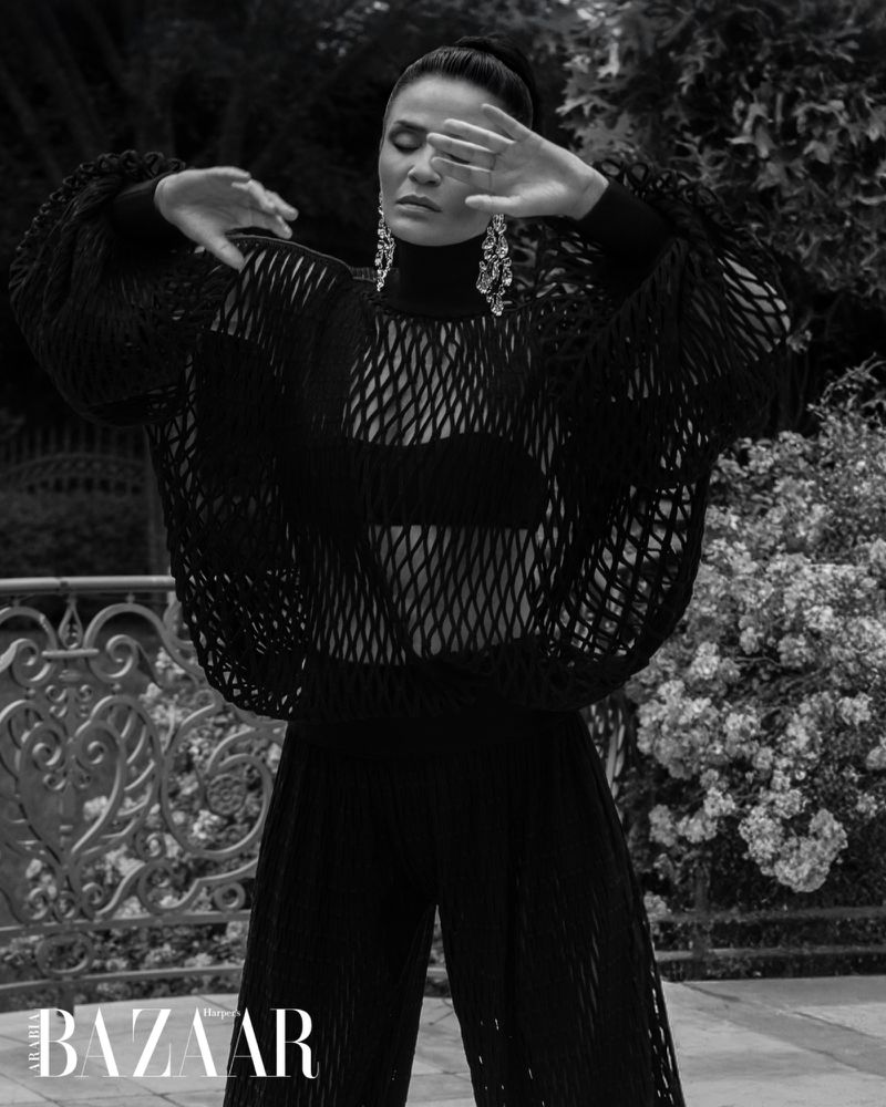 Monochrome fashion portrait: adult female in net top, wide pants, chandelier earrings, hand gesture, outdoor setting, 'Harper's Bazaar' logo.