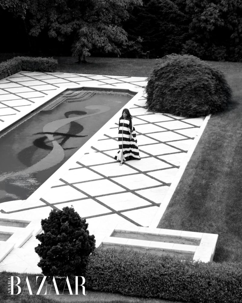 Elegant woman in striped dress walks by outdoor pool in a luxury garden setting, black and white photography, Harper's Bazaar logo.