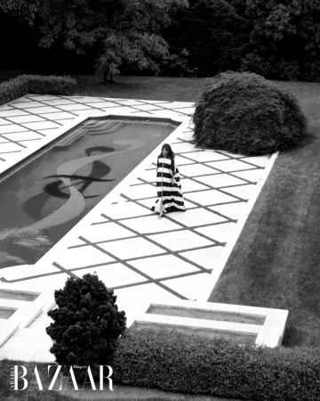 Elegant woman in striped dress walks by outdoor pool in a luxury garden setting, black and white photography, Harper's Bazaar logo.