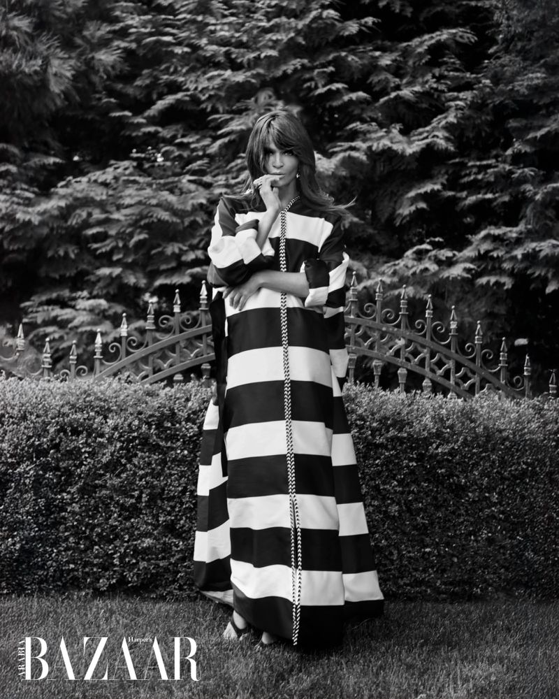 Black and white fashion shot of a woman in a striped gown standing in a garden with a decorative gate and lush green foliage.