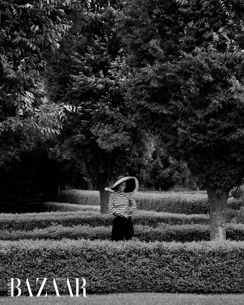 Woman in a striped shirt and wide-brimmed hat stands in a garden maze with trees, captured in black and white, Bazaar logo at the bottom.