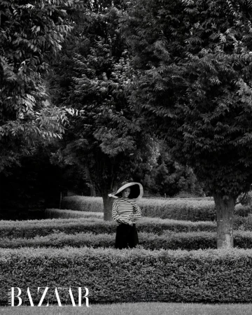 Woman in a striped shirt and wide-brimmed hat stands in a garden maze with trees, captured in black and white, Bazaar logo at the bottom.