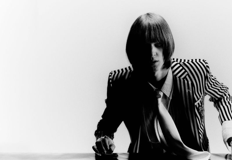 Black and white portrait of a male model wearing a striped suit and tie, seated at a table, with a bright white background.
