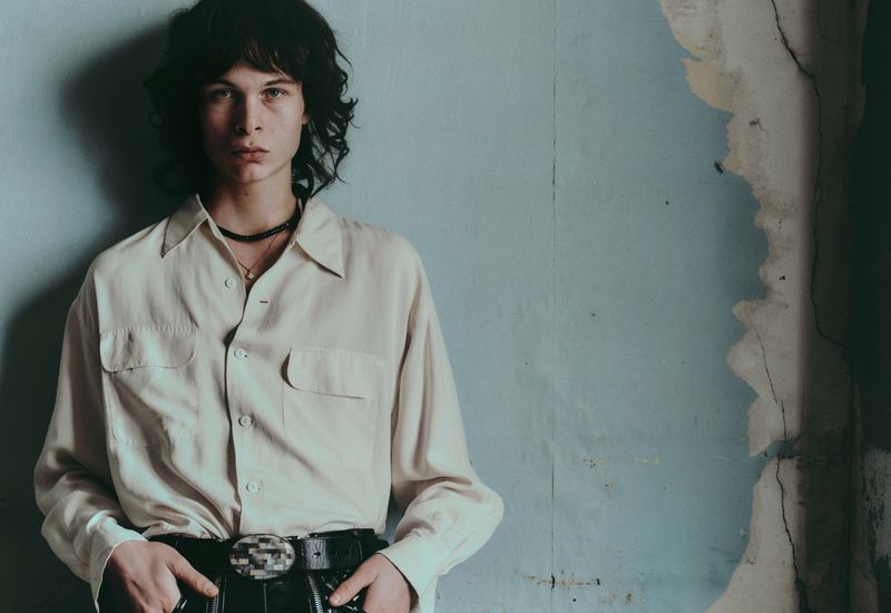 Model Mats Chabot in an off-white button-down shirt and black belt leaning against a distressed blue wall with peeling plaster.