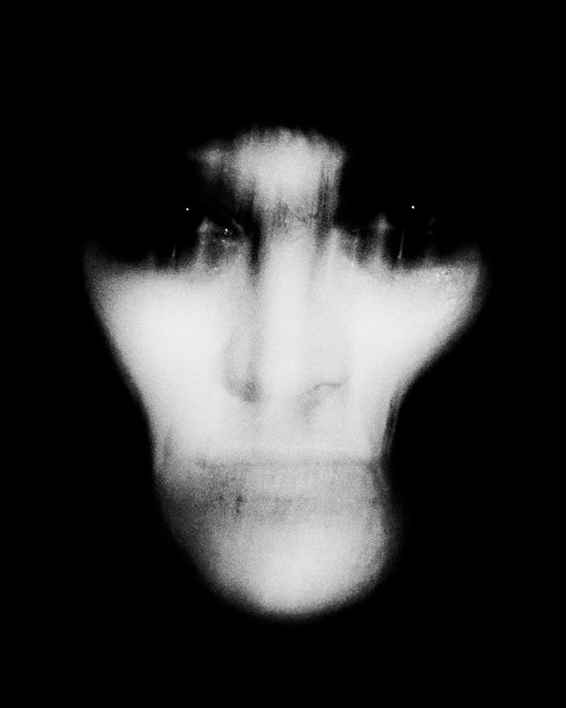 Monochromatic portrait of an adult with blurred facial features in a high-contrast, high-key composition against a stark black background.