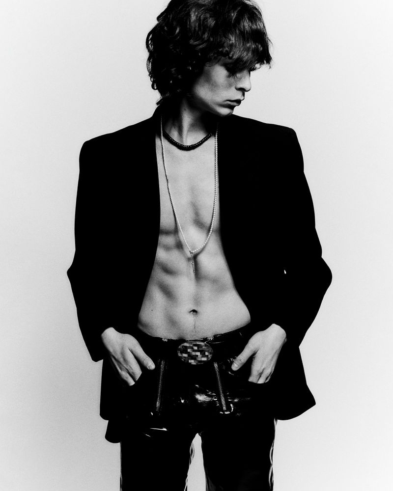 Black and white portrait of a stylish adult male wearing a black blazer and pants, showcasing his torso and necklaces in a studio.
