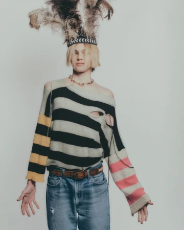 Fashion shot of a fair-skinned person with blond hair wearing a striped sweater, jeans, and feathered headpiece against a white backdrop.
