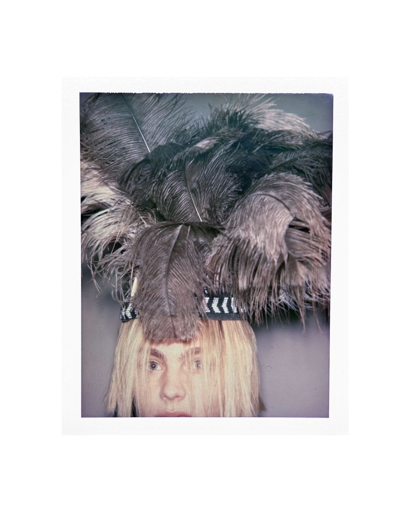 Person wearing a large feathery headdress with blond hair partially covering their face in a studio shot with grey background.
