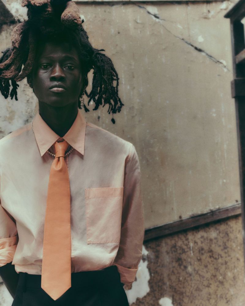 Portrait of a young Black man with dreadlocks wearing a light pink collared shirt and peach-colored tie in an outdoor setting.