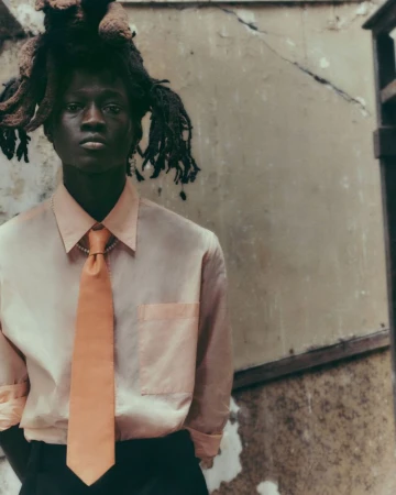 Portrait of a young Black man with dreadlocks wearing a light pink collared shirt and peach-colored tie in an outdoor setting.