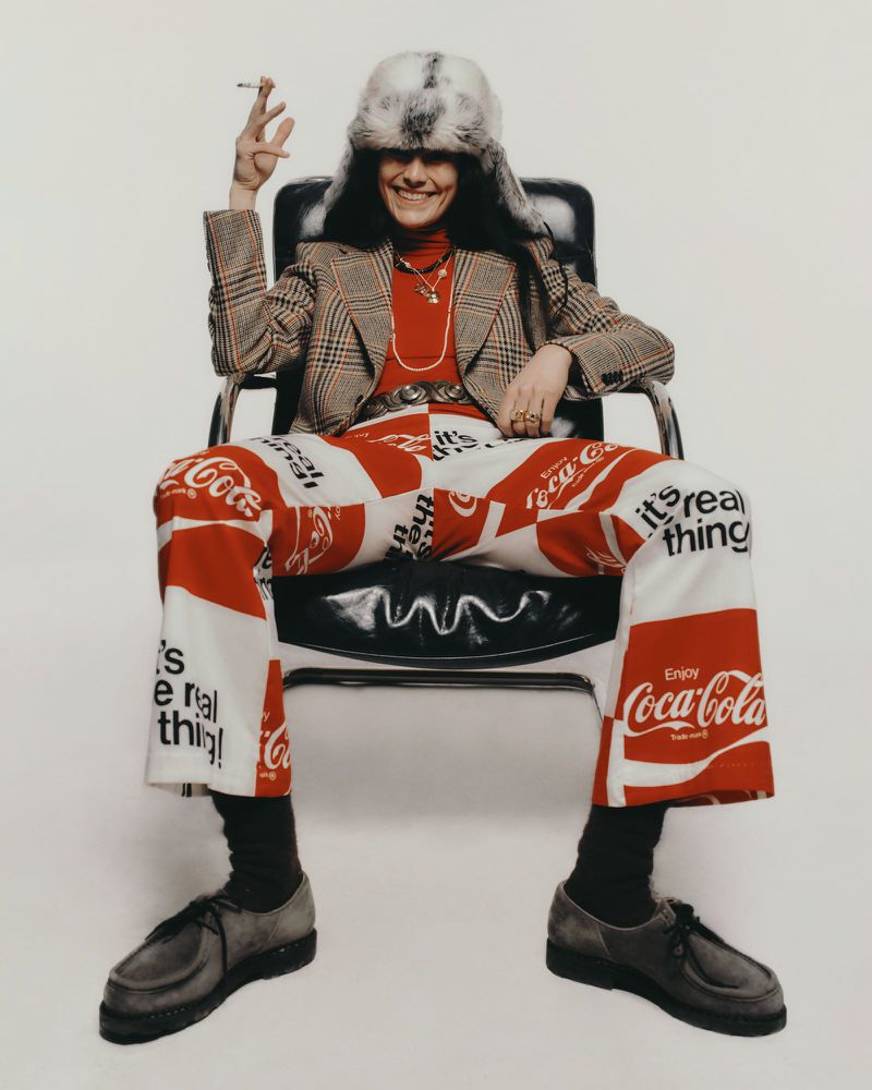 Person in Coca-Cola pants and a fur hat seated in a chair, holding a cigarette against a white backdrop in a studio setting.