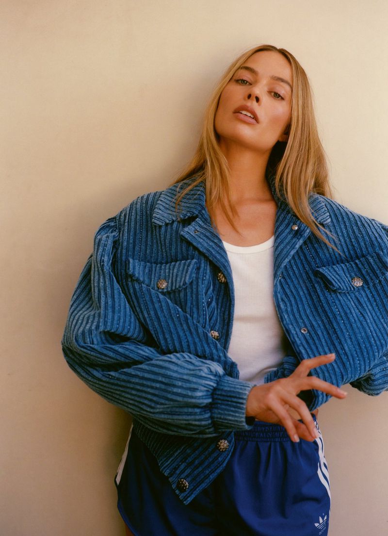 Blonde woman in blue corduroy jacket and blue track shorts poses against a neutral wall in soft, diffused lighting.
