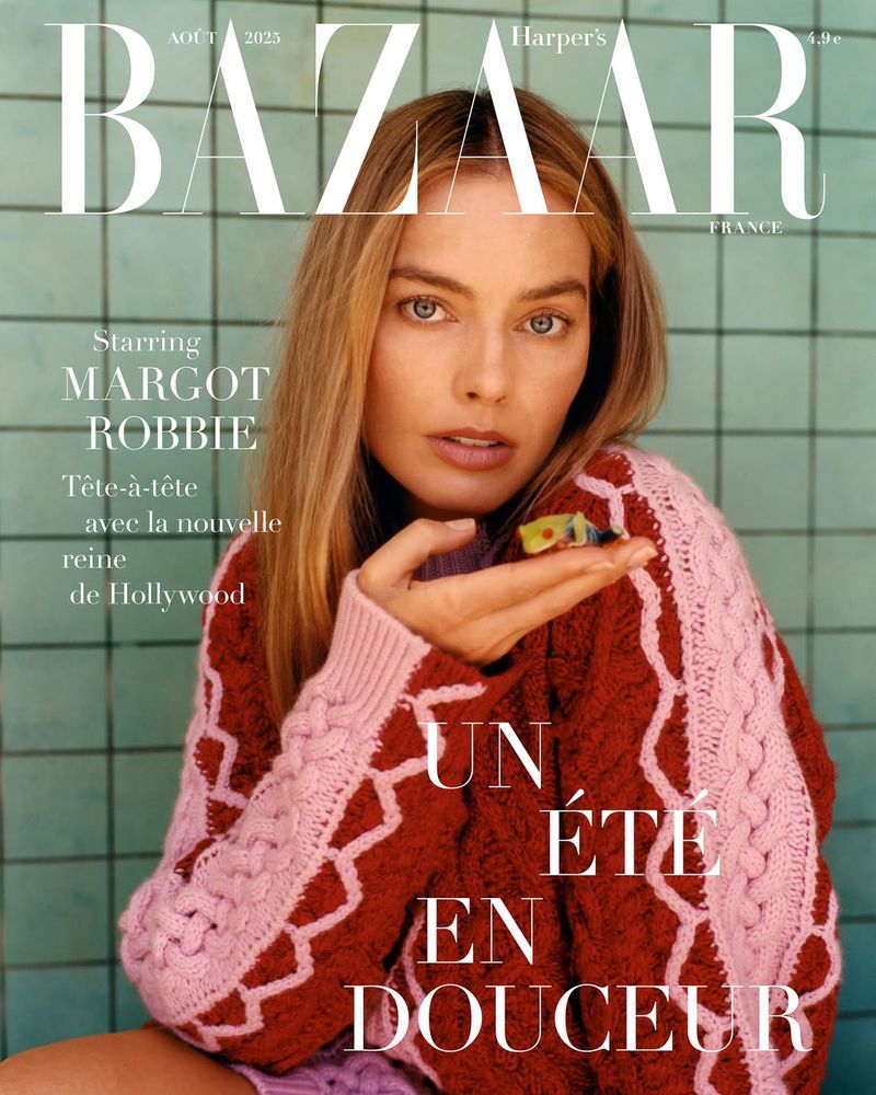 Magazine cover featuring Margot Robbie with a toy figure, wearing a pink and burgundy cable-knit sweater against a tiled backdrop.