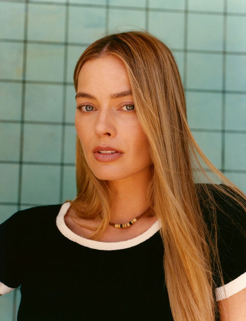Close-up studio portrait of a blonde woman with light skin wearing a black shirt and necklace, gazing at the camera against a teal tiled wall.
