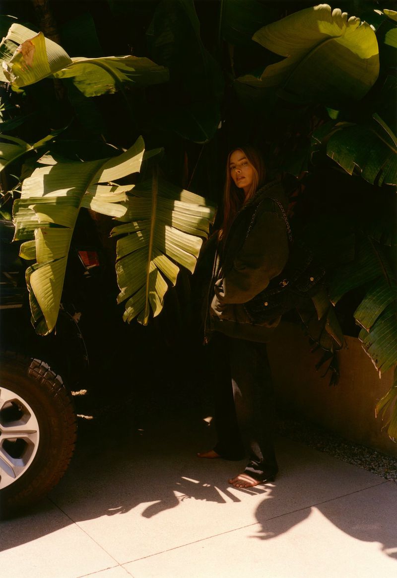 A woman stands by a car tire amidst lush tropical foliage and warm sunlight, wearing casual attire, photographed in an outdoor setting.