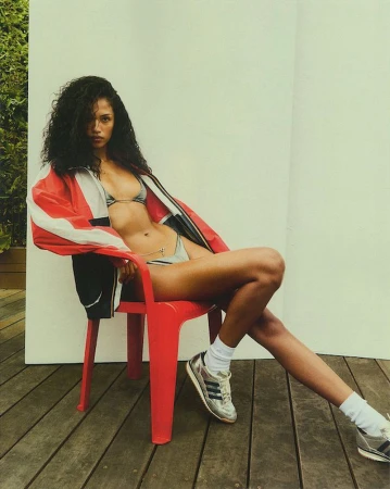 A woman with dark, curly hair, wearing a bikini top and a red and white jacket, sits on a red chair on a wooden deck.