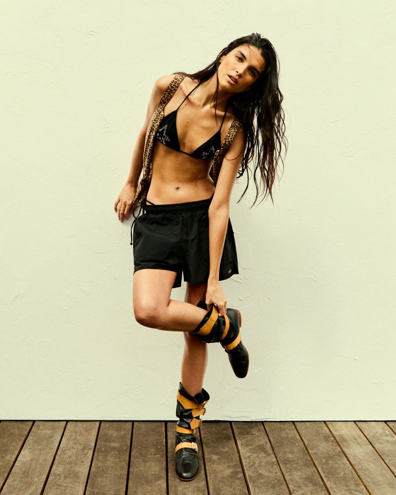 Brunette woman in a black bikini top, leopard print vest, black shorts, and high boots poses in front of a white wall and wood floor.
