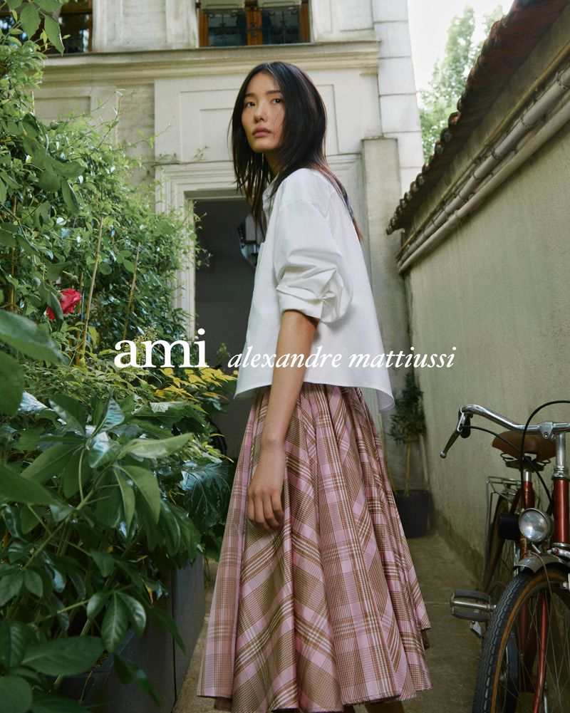 A person models a white shirt and plaid skirt in a relaxed outdoor setting, with a bicycle and greenery in the background.