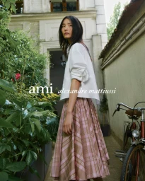 Model wearing a white shirt and pleated pink plaid skirt outdoors by a bicycle and garden foliage.