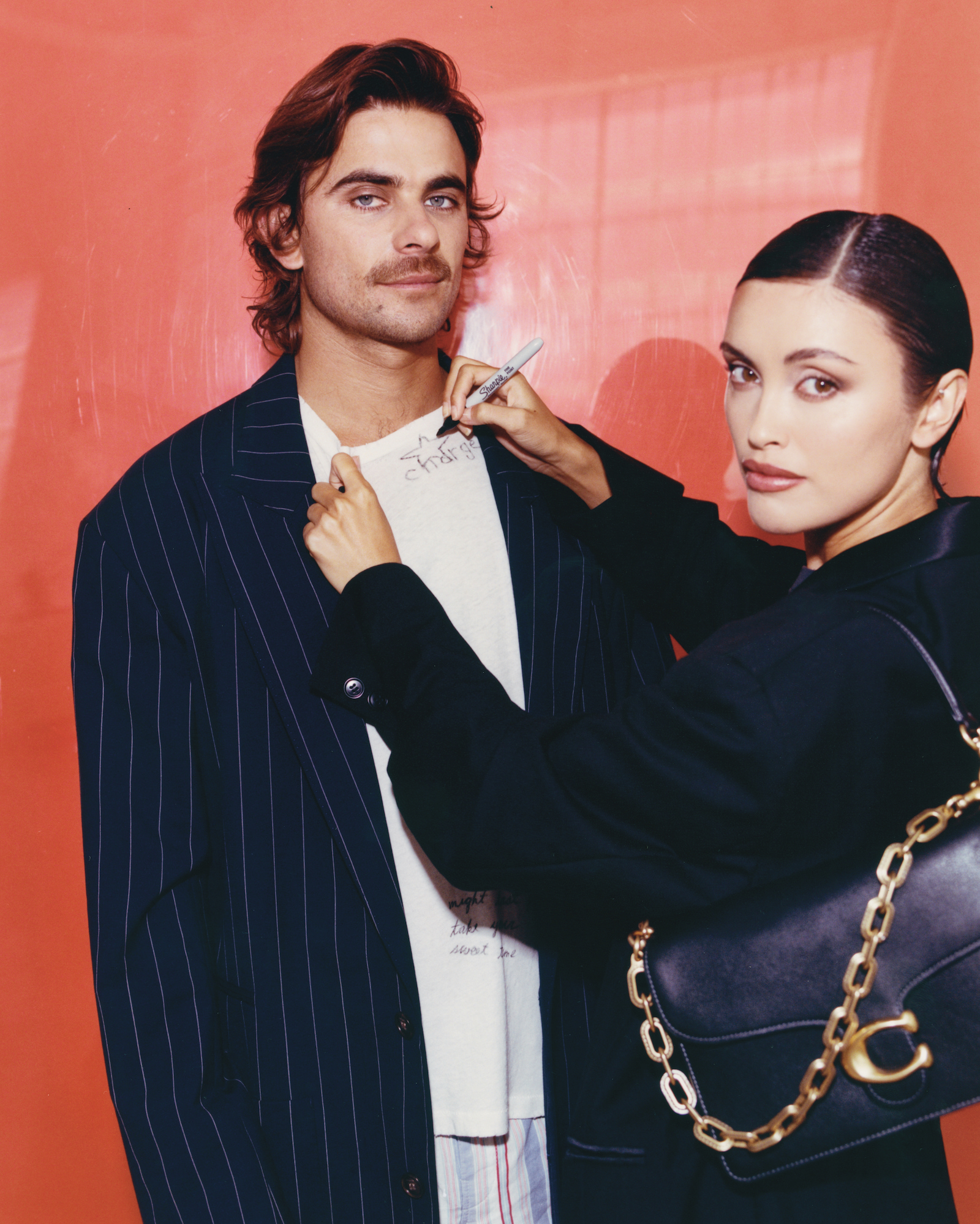 Man and woman pose indoors with a black purse; the woman is holding a pen, with an orange gradient background, and soft lighting.