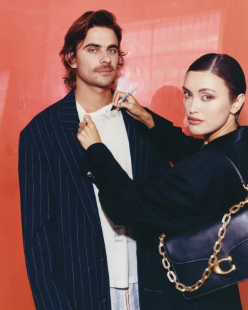 Man and woman pose indoors with a black purse; the woman is holding a pen, with an orange gradient background, and soft lighting.