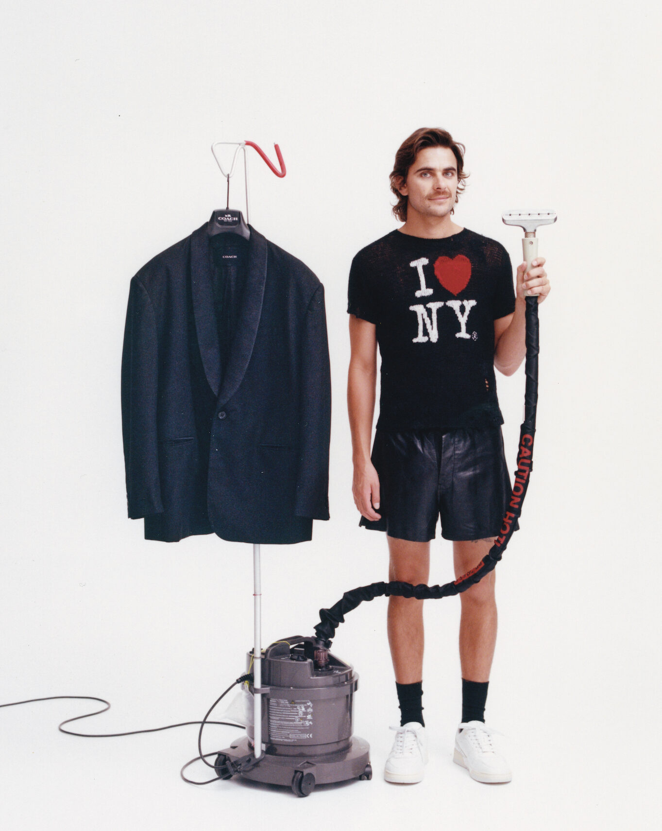 Man stands next to a steamer, wearing an 'I love NY' shirt, black shorts, and white shoes, posed in front of a white backdrop.