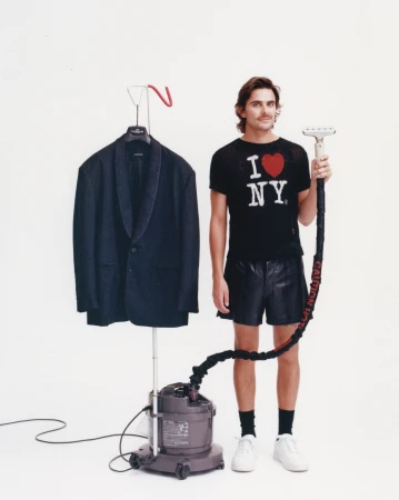Man stands next to a steamer, wearing an 'I love NY' shirt, black shorts, and white shoes, posed in front of a white backdrop.