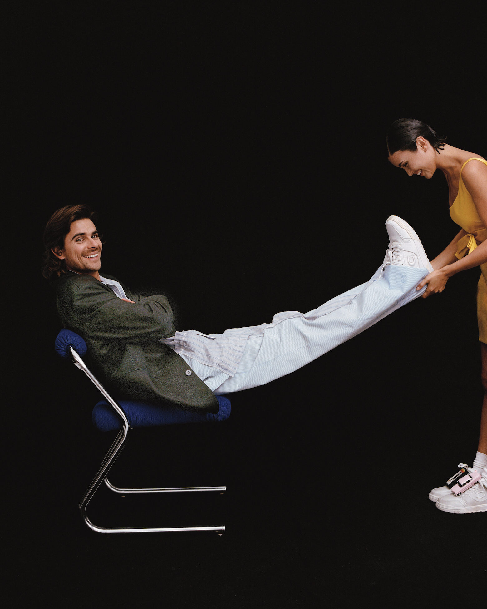 Man seated on a blue chair with legs extended upward while a woman holds his shoes against a black background.