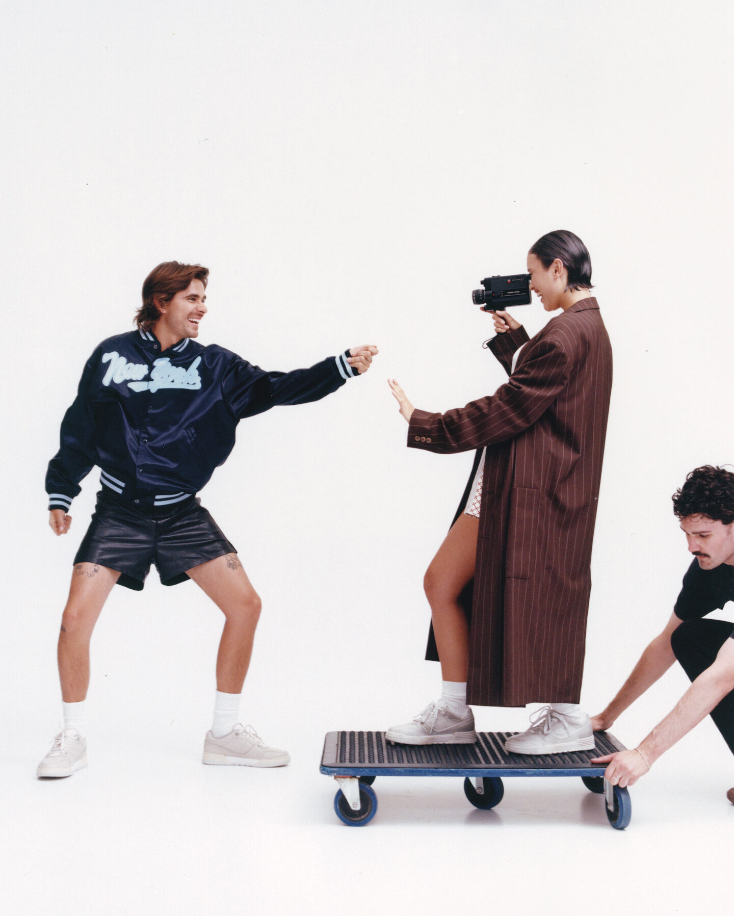 Three people in a white studio. One in athletic wear reaching, a woman in a coat filming, and another pushing them on a dolly.