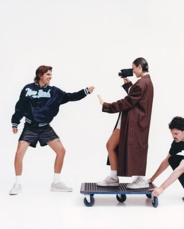 Three people in a white studio. One in athletic wear reaching, a woman in a coat filming, and another pushing them on a dolly.